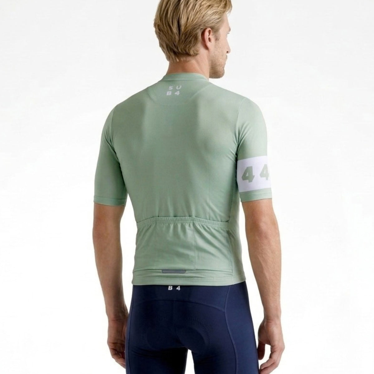 A man wearing a light sage green short-sleeve cycling jersey from the back, paired with dark navy cycling shorts, against a plain white background.