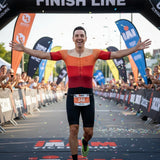 Athlete crossing the finish line in a triathlon with confetti and spectators in the background.